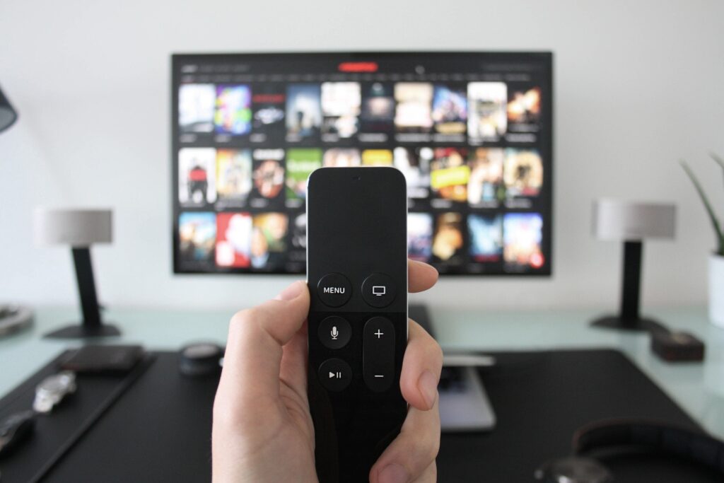 Una mano sosteniendo un control remoto moderno (menos opciones en el mando). De fondo se ve difuso un televisor con imágenes de programas de tv a elegir. 